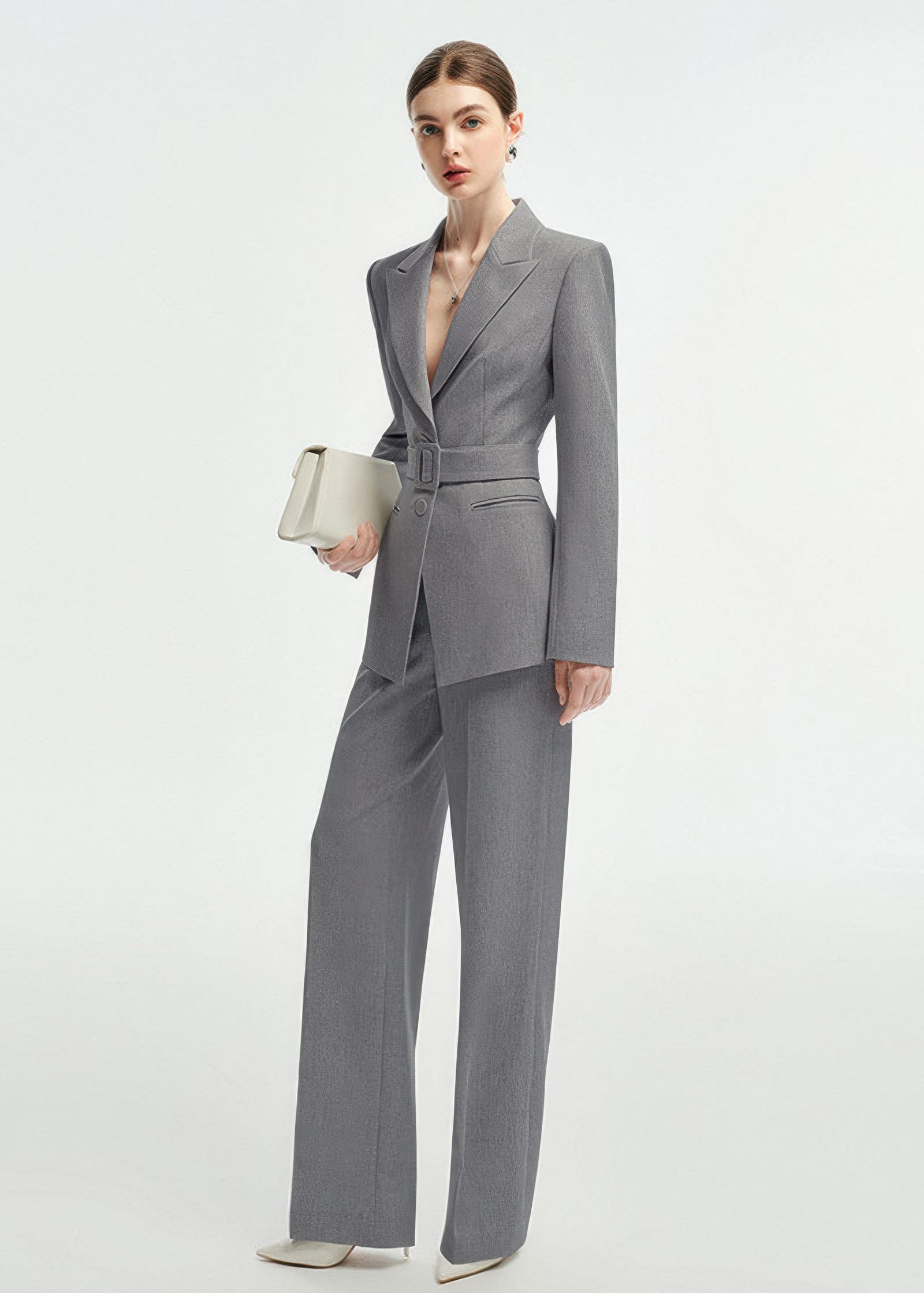Three-quarter length shot of a woman in a grey belted blazer and matching trousers, holding a white clutch bag.