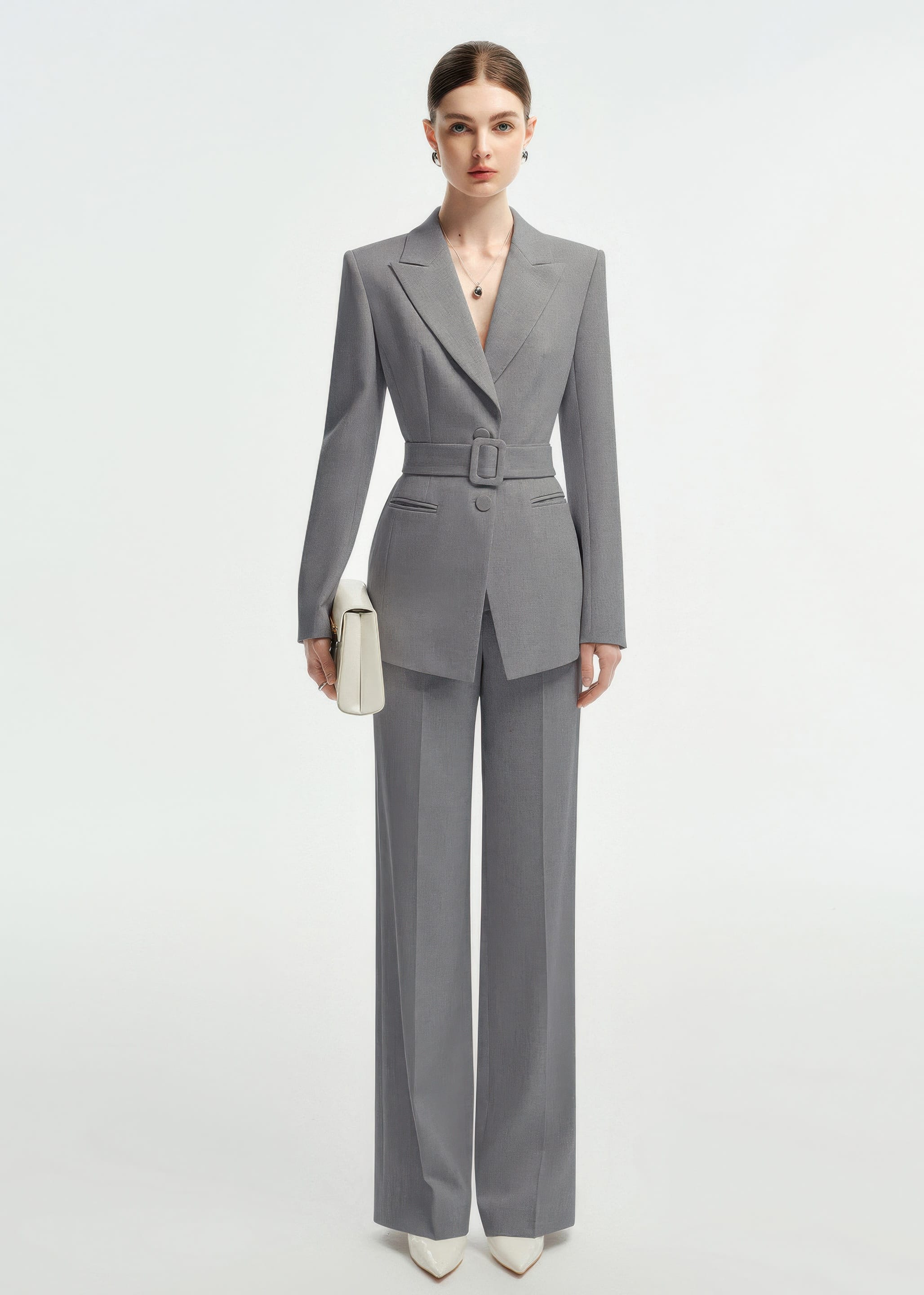 Full-length front view of a woman posing in a professional grey belted suit with white pointed-toe heels.