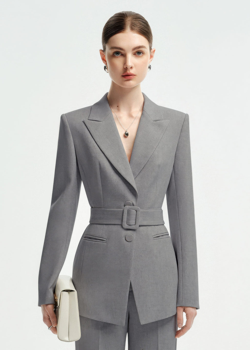 Close-up portrait of a woman wearing a grey belted suit jacket with sharp lapels and a silver pendant necklace.