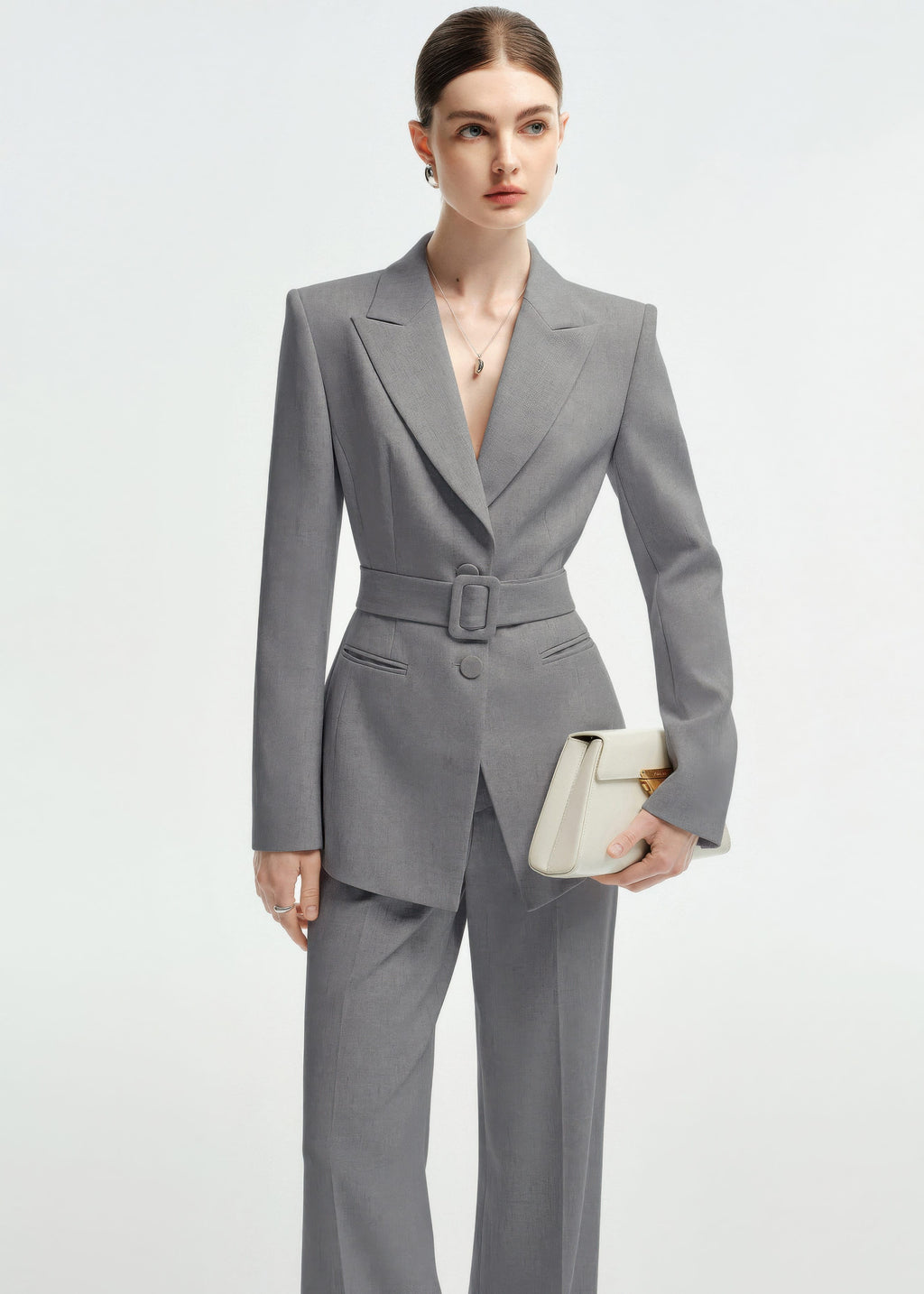 Medium shot of a woman in the belted grey blazer, holding a structured cream-colored clutch.