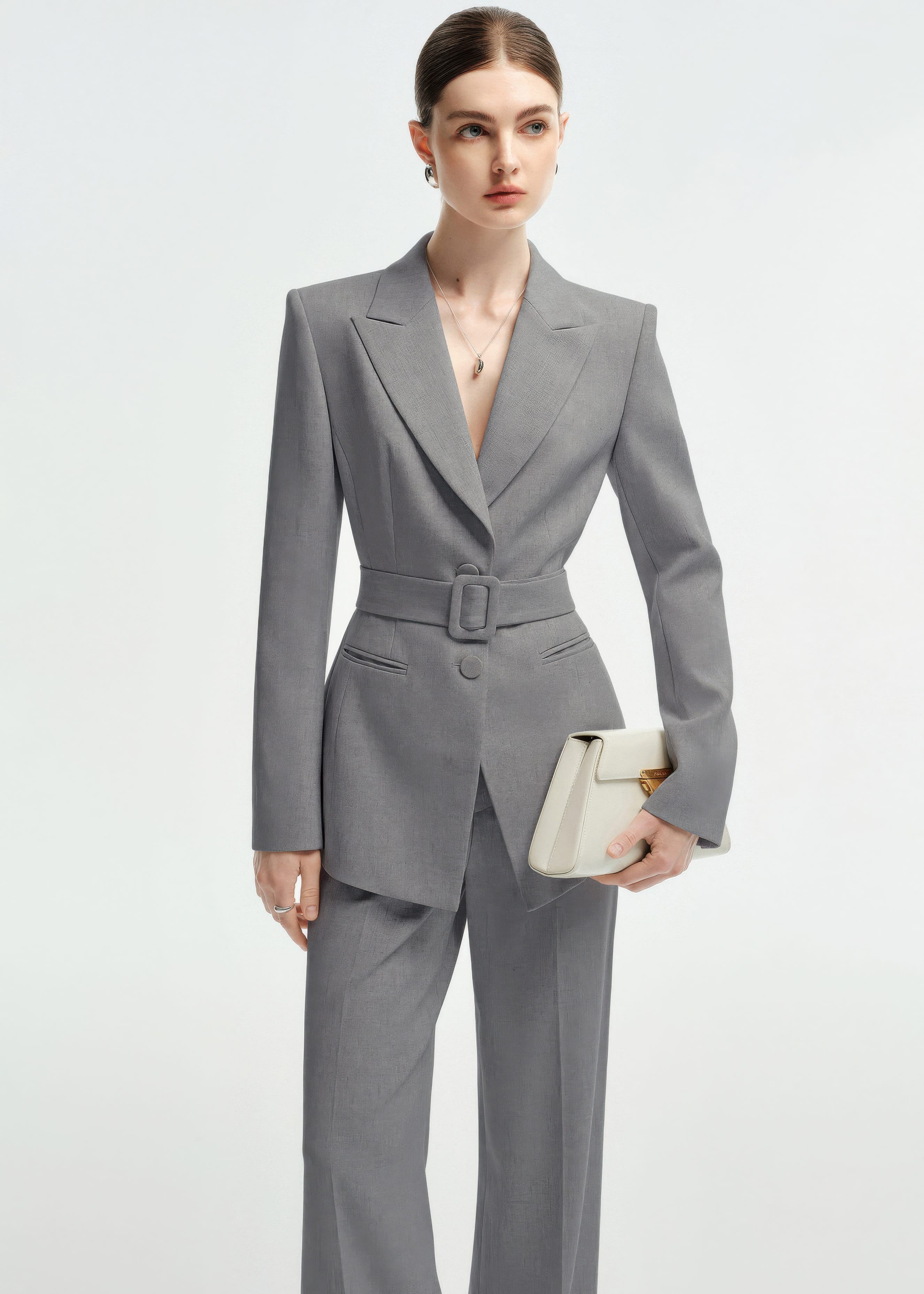 Medium shot of a woman in the belted grey blazer, holding a structured cream-colored clutch.