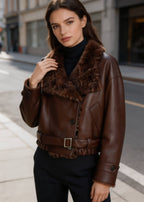 Front view of a woman wearing a dark brown faux leather aviator jacket with a plush fur collar on a city street.