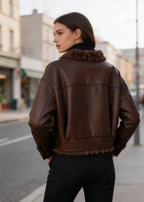 Rear angle of a female model wearing a brown leather winter jacket with fur-trimmed hem outdoors.