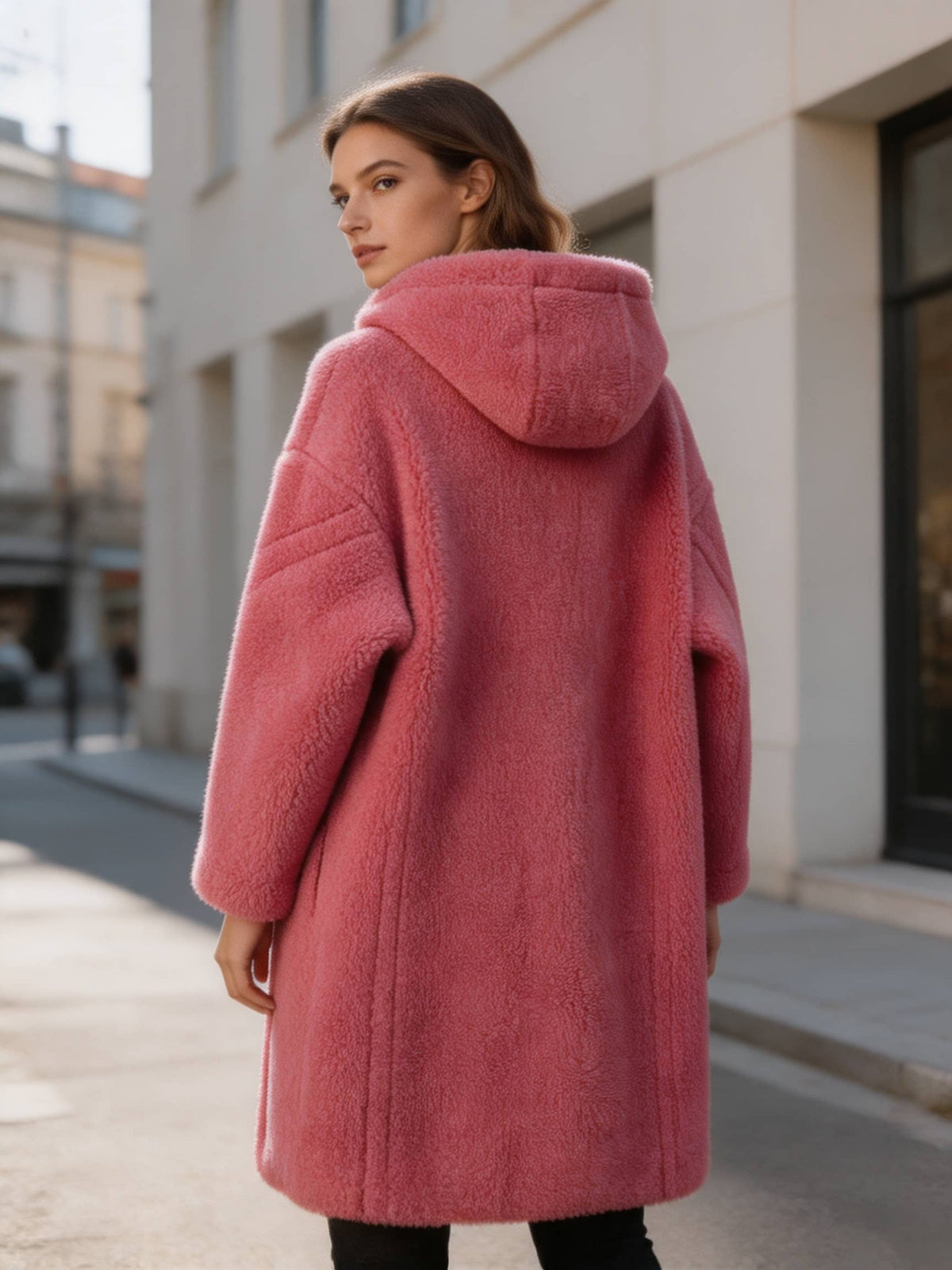 Rear view shot of a woman wearing a long fuchsia pink plush shearling coat with a hood on a city street.