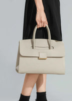 A close-up lifestyle shot focusing on the hand holding the tote, emphasizing the bag's elegant design and the contrast between the cream leather and a black dress.