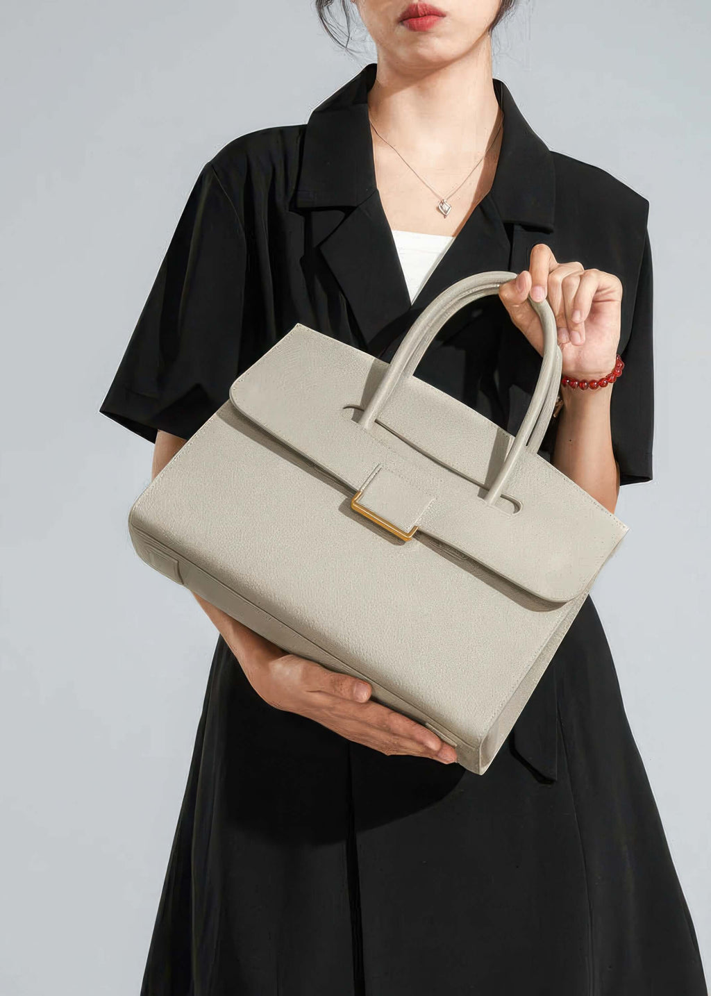 A lifestyle image of a woman holding the tote handbag with both hands in front of her, showcasing the bag as a statement accessory against a dark outfit.