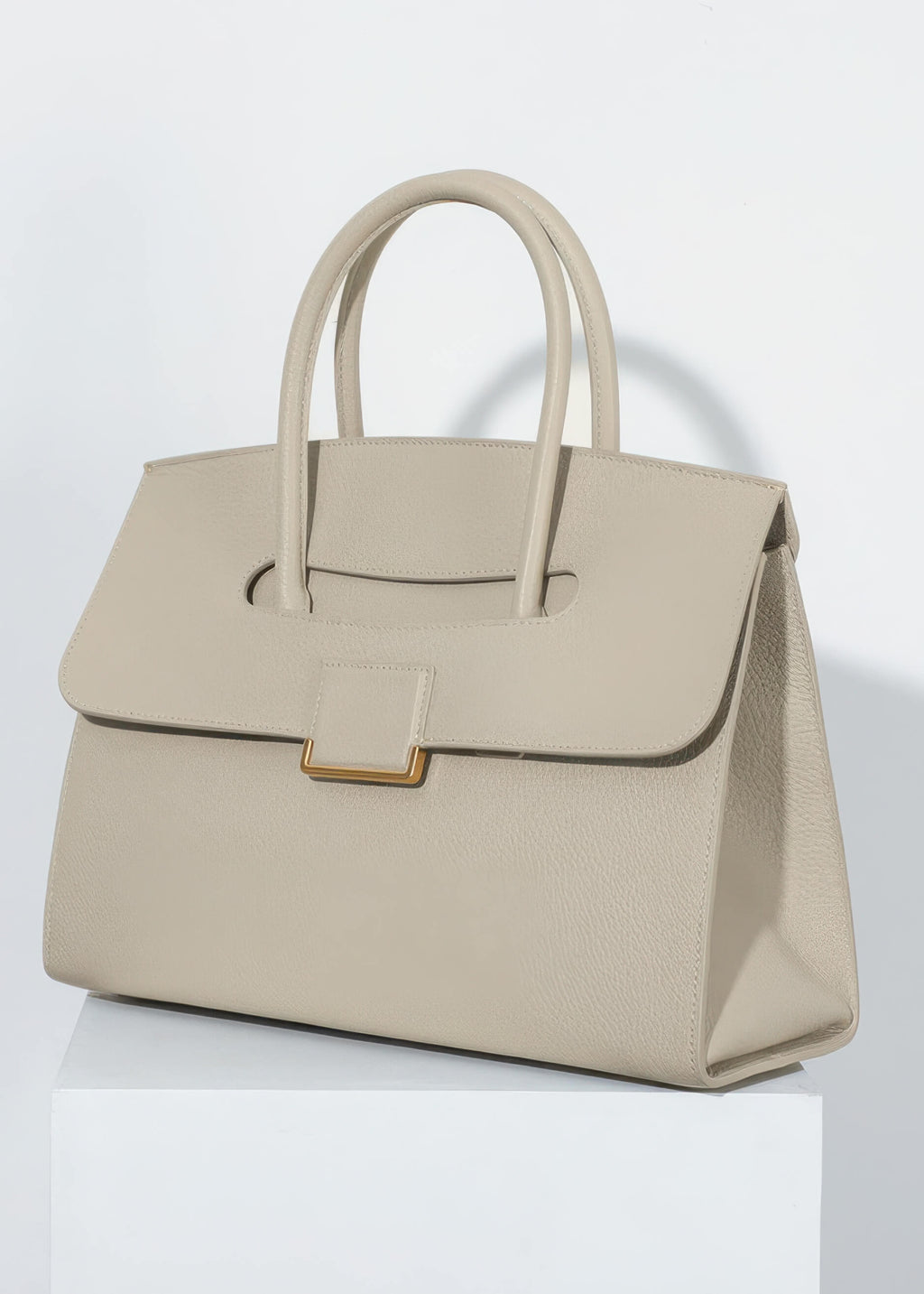 A professional studio shot of the genuine leather tote sitting on a white pedestal. The lighting highlights the fine pebbled texture of the leather and the gold-tone hardware.