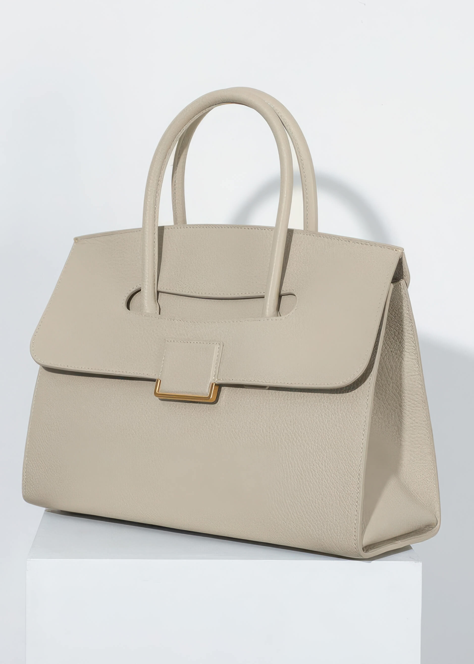 A professional studio shot of the genuine leather tote sitting on a white pedestal. The lighting highlights the fine pebbled texture of the leather and the gold-tone hardware.