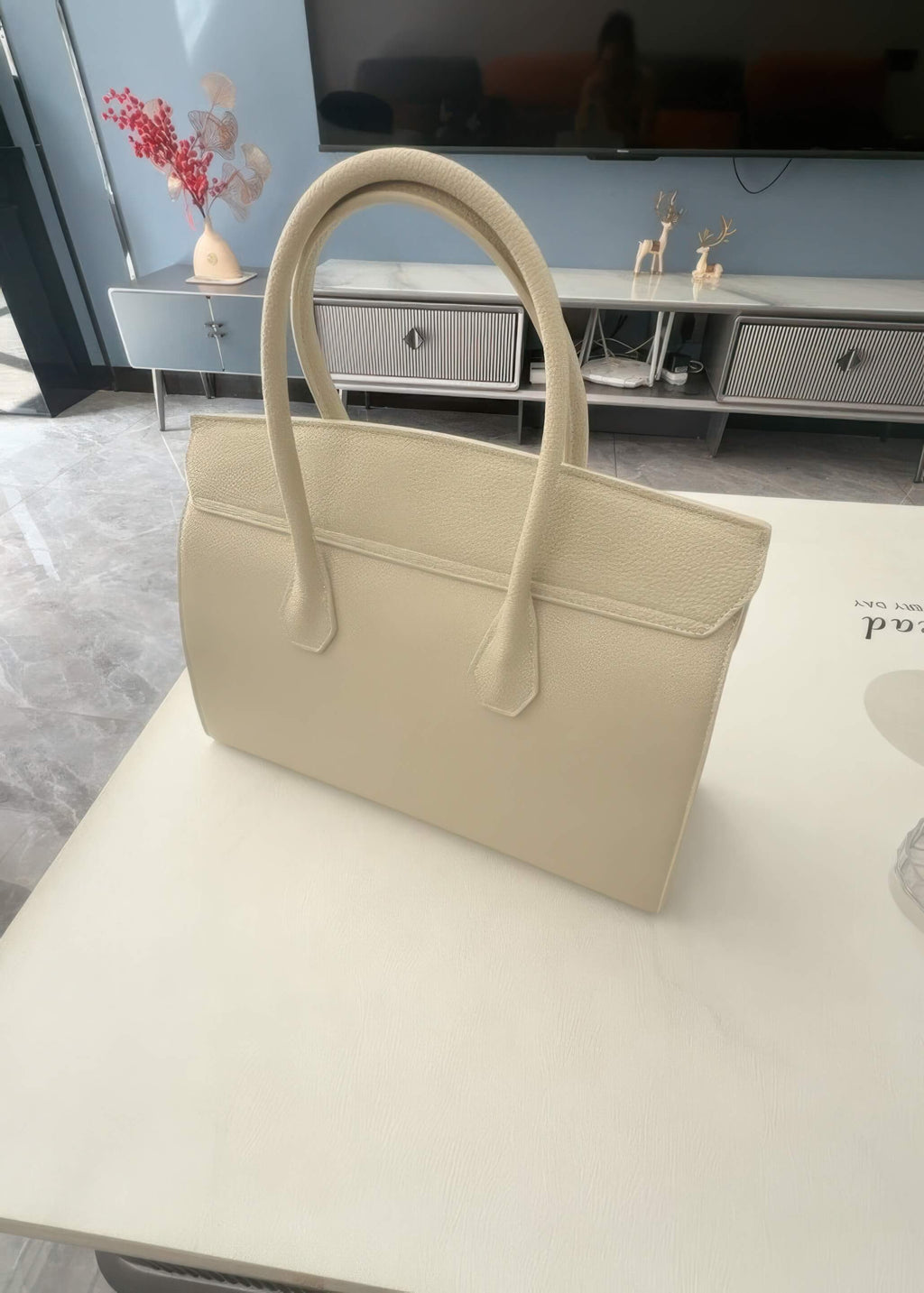 The back view of the cream-colored leather tote handbag, showing the smooth attachment points of the dual handles and the clean, structured silhouette against a home interior background.