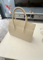 The back view of the cream-colored leather tote handbag, showing the smooth attachment points of the dual handles and the clean, structured silhouette against a home interior background.