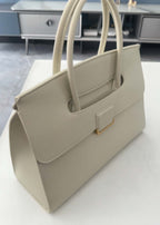 A high-angle, close-up shot of a cream-colored, textured genuine leather tote handbag. It features a structured rectangular shape, dual rolled top handles, and a unique flap closure with a gold-tone rectangular accent.