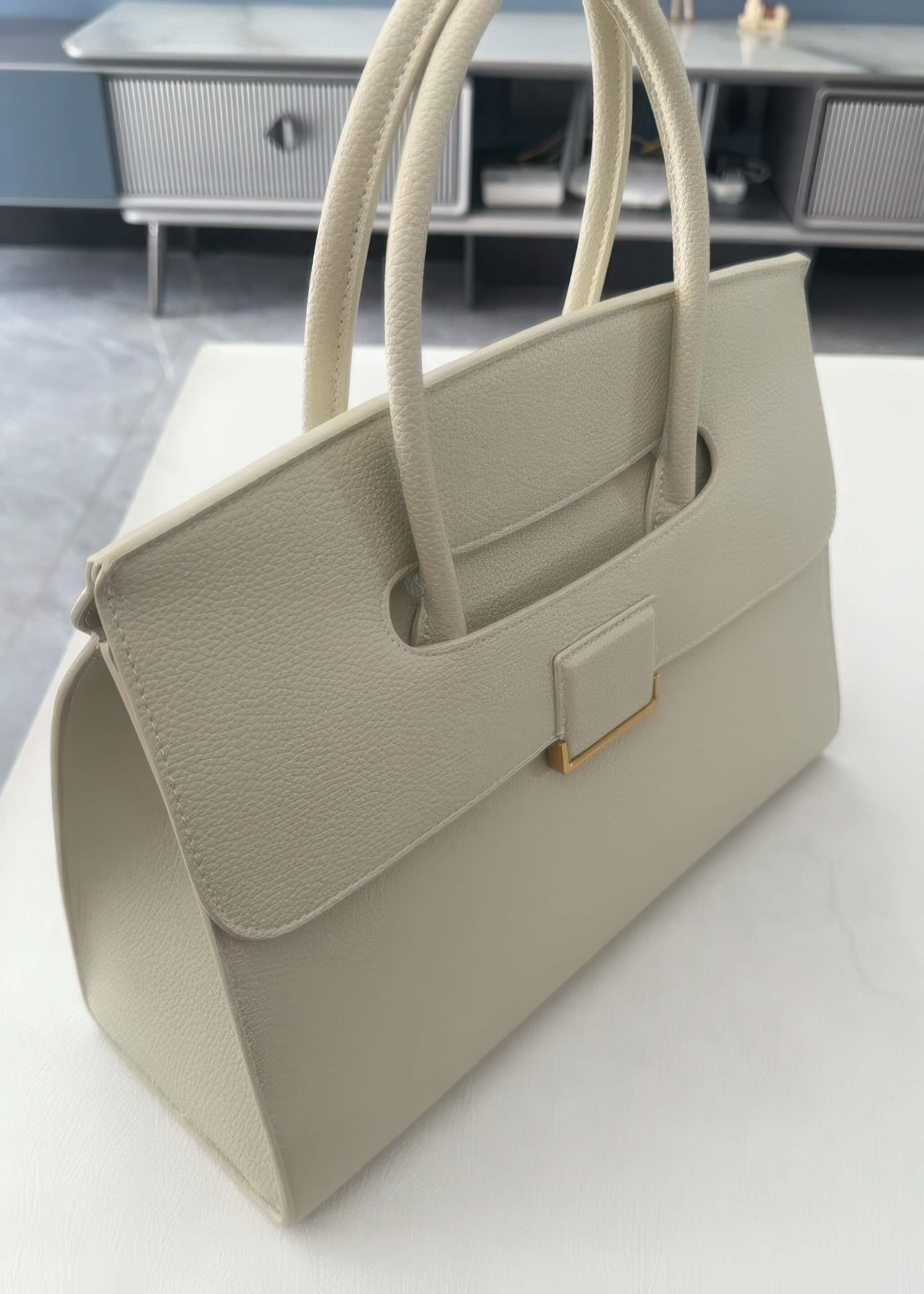 A high-angle, close-up shot of a cream-colored, textured genuine leather tote handbag. It features a structured rectangular shape, dual rolled top handles, and a unique flap closure with a gold-tone rectangular accent.