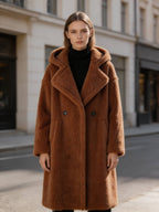 Model wearing a brown long hooded shearling wool teddy coat standing outdoors on a city street