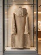 Back view display of a women's beige hooded wool shearling long teddy coat hanging in a boutique window showcase.