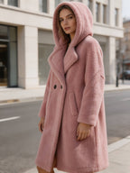 Three-quarter view of a female model wearing a cozy pink hooded shearling teddy coat while walking down a city street.