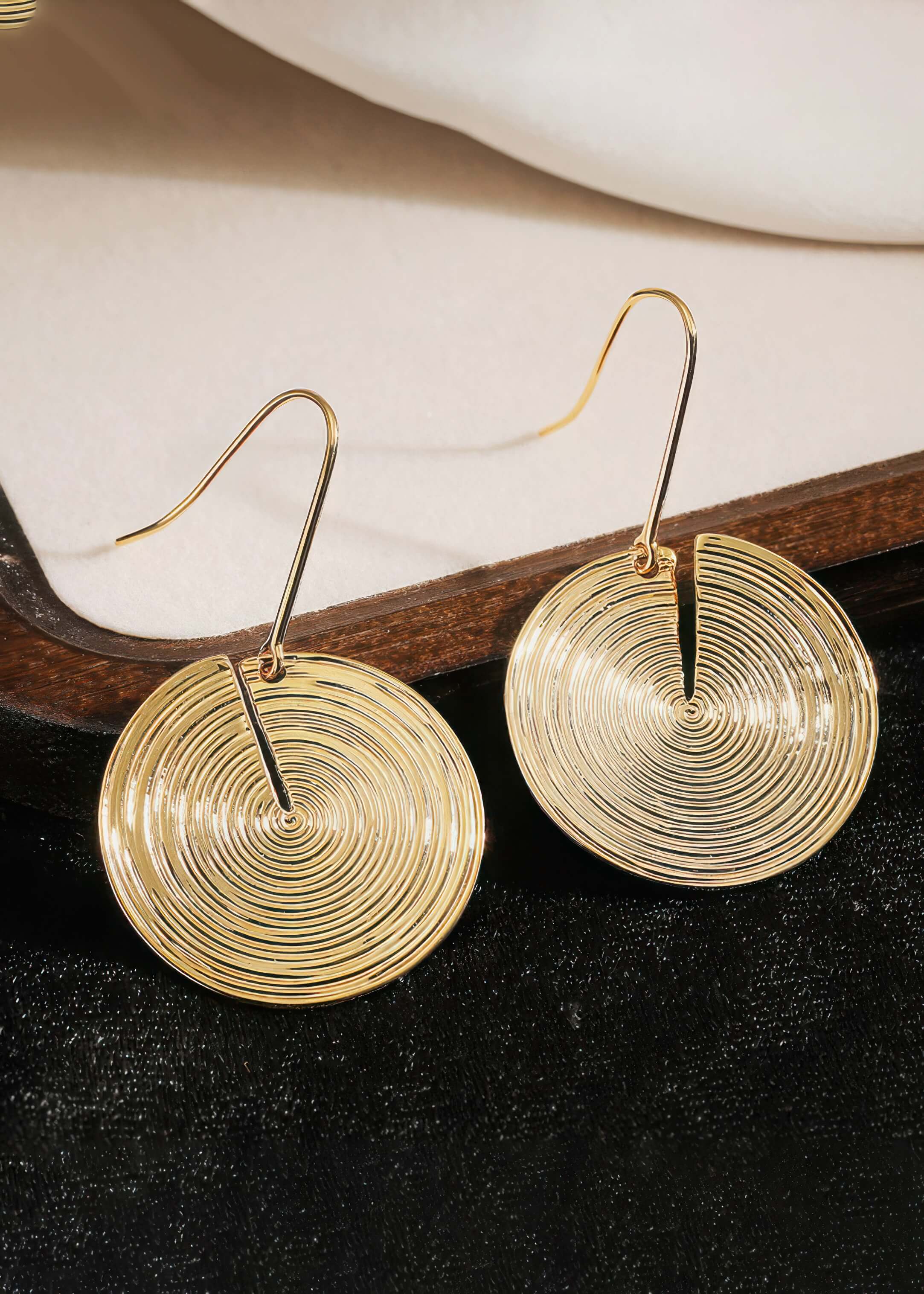 A pair of gold concentric circle drop earrings laying on a light-colored surface with dark wood accents.