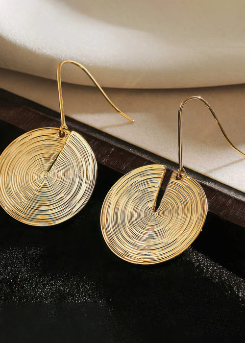 Close-up of gold metal drop earrings with a concentric circle design, resting on a dark, textured background.