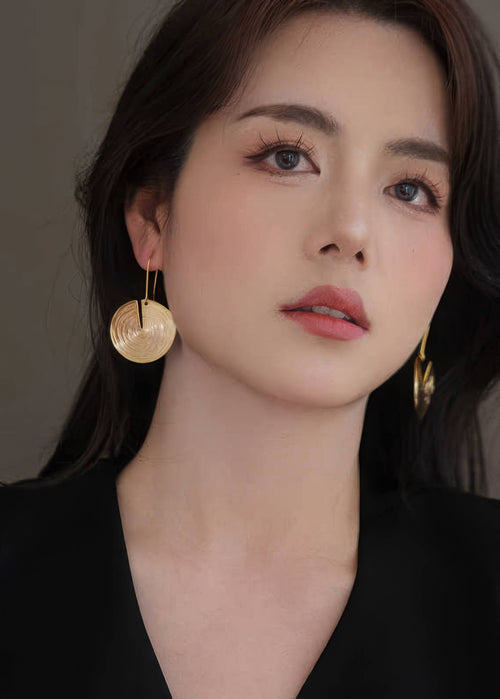Front-facing portrait of a woman wearing gold-toned metal drop earrings with a concentric circular "tree ring" texture.