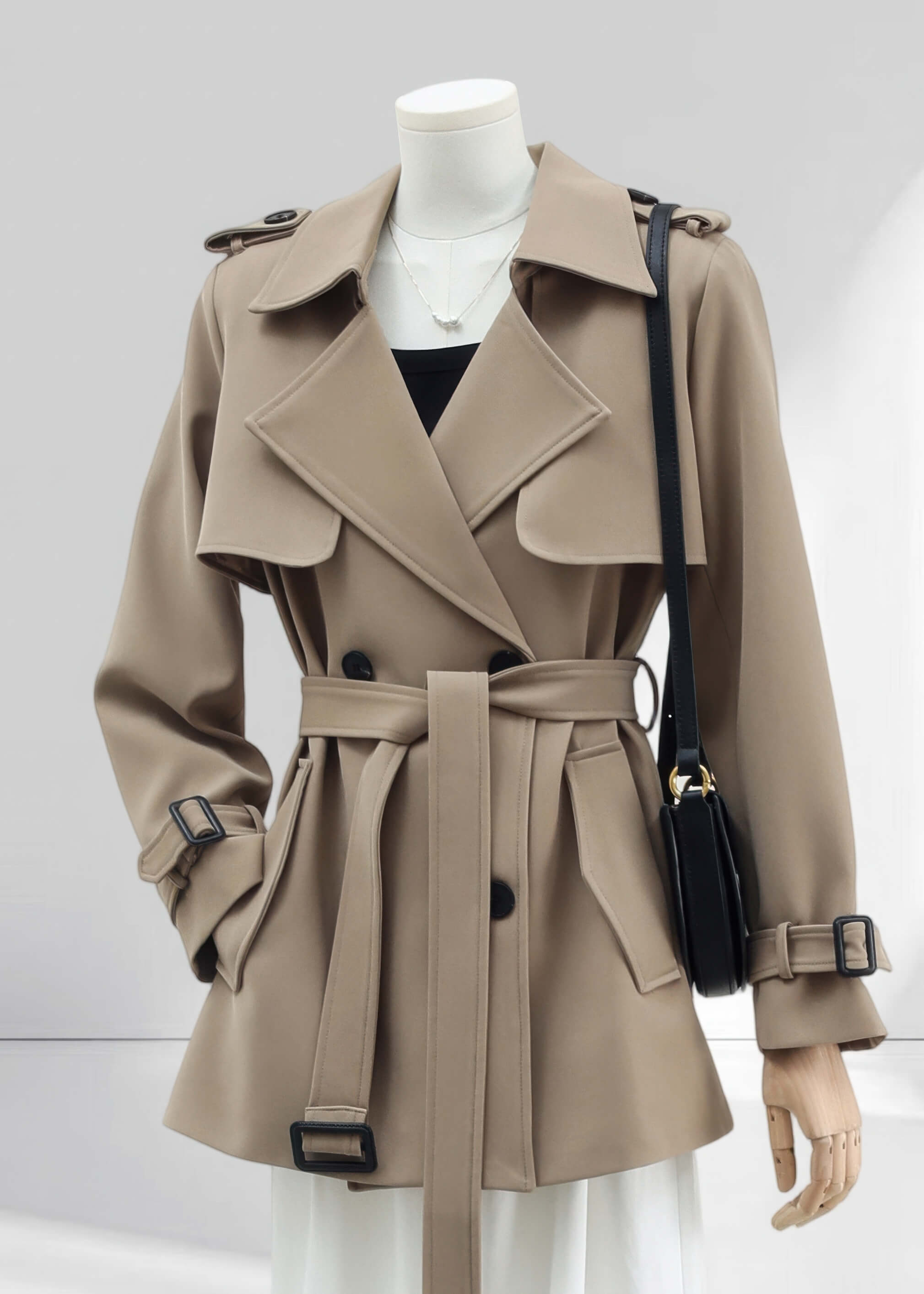 A front-facing, medium shot of the khaki short trench coat. It shows the coat styled with a black top and a delicate pearl necklace, featuring a structured storm flap and a matching fabric belt tied in the center.
