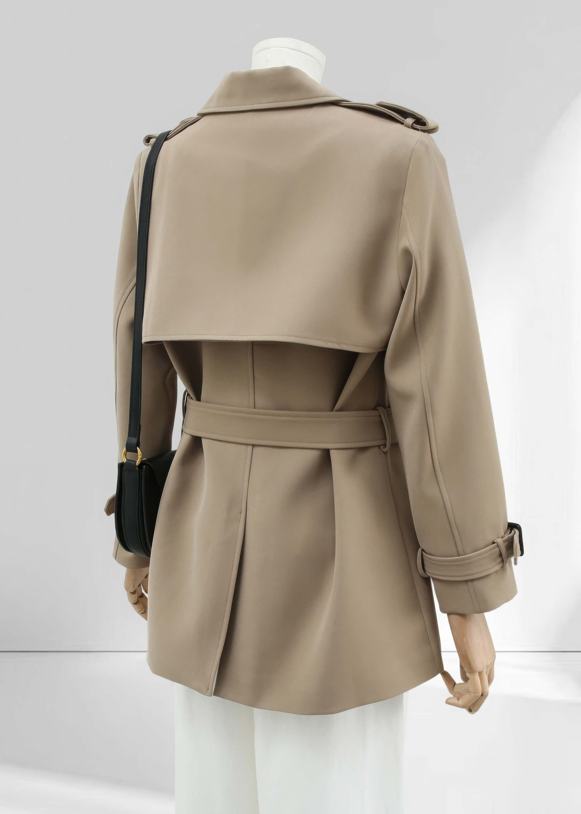A back view of the short trench coat on the mannequin. This shot emphasizes the large storm flap across the shoulders, the central back vent, and the clean silhouette of the belted waist.