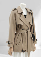 A side-angle view of a khaki short trench coat on a mannequin. The image highlights the double-breasted lapels, a tied waist belt, and adjustable cuff straps with black buckles.