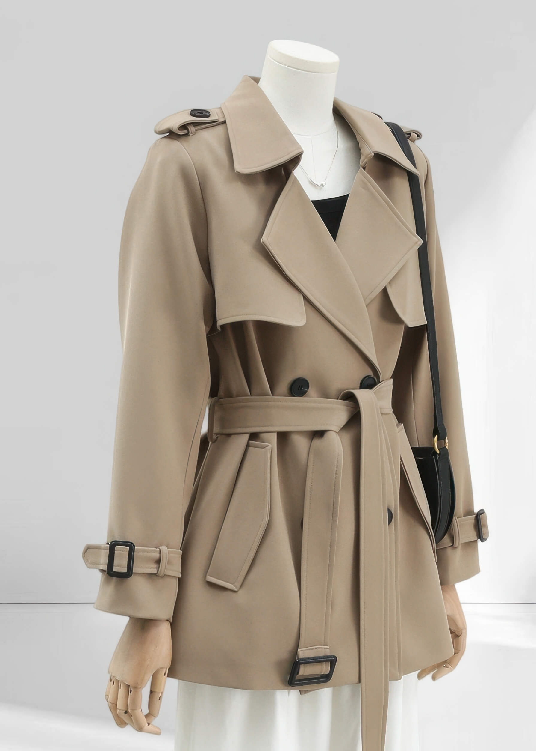 A side-angle view of a khaki short trench coat on a mannequin. The image highlights the double-breasted lapels, a tied waist belt, and adjustable cuff straps with black buckles.