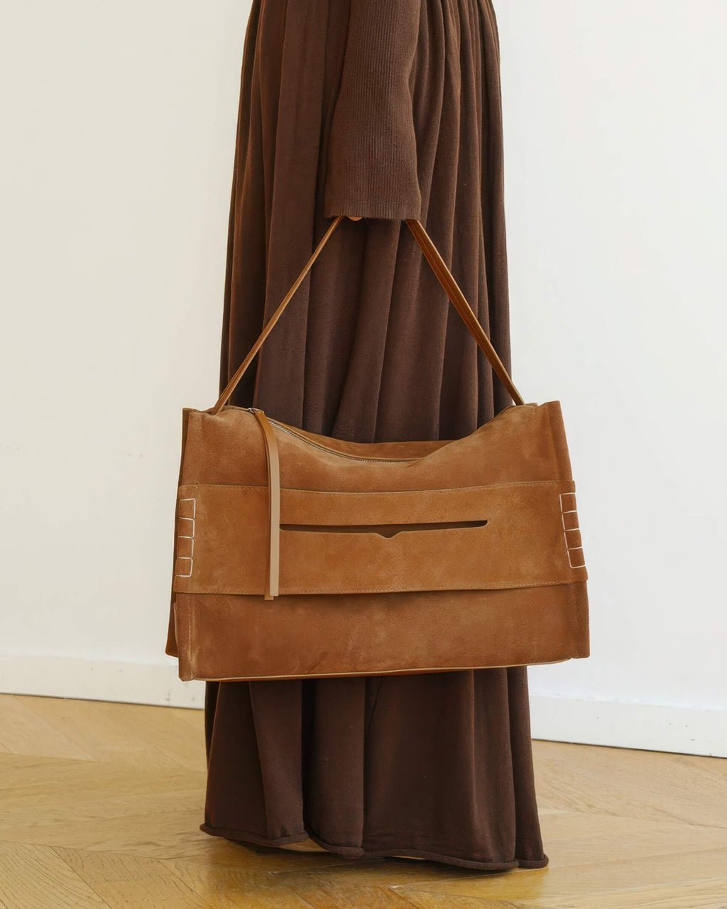 Full-length lifestyle view of a woman carrying a minimalist large tan suede shoulder bag, showing its rectangular silhouette and long strap.