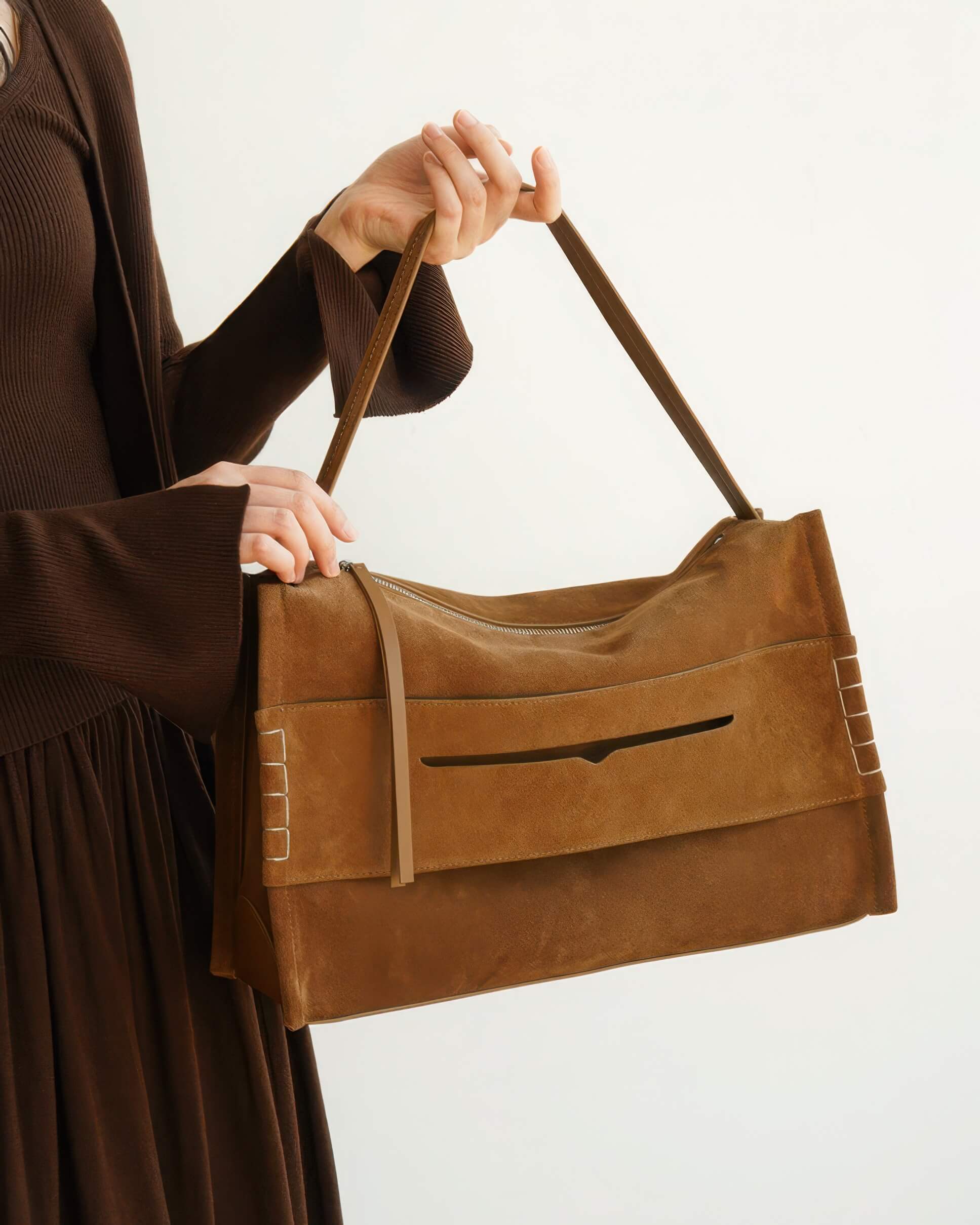 Close-up of a person holding a large brown suede shoulder bag with a unique horizontal slit pocket and white contrast stitching on the sides.