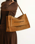 A side-view lifestyle shot of a woman wearing the large brown suede shoulder bag, showcasing its soft rectangular structure, side whipstitch details, and the unique slit-style front pocket.