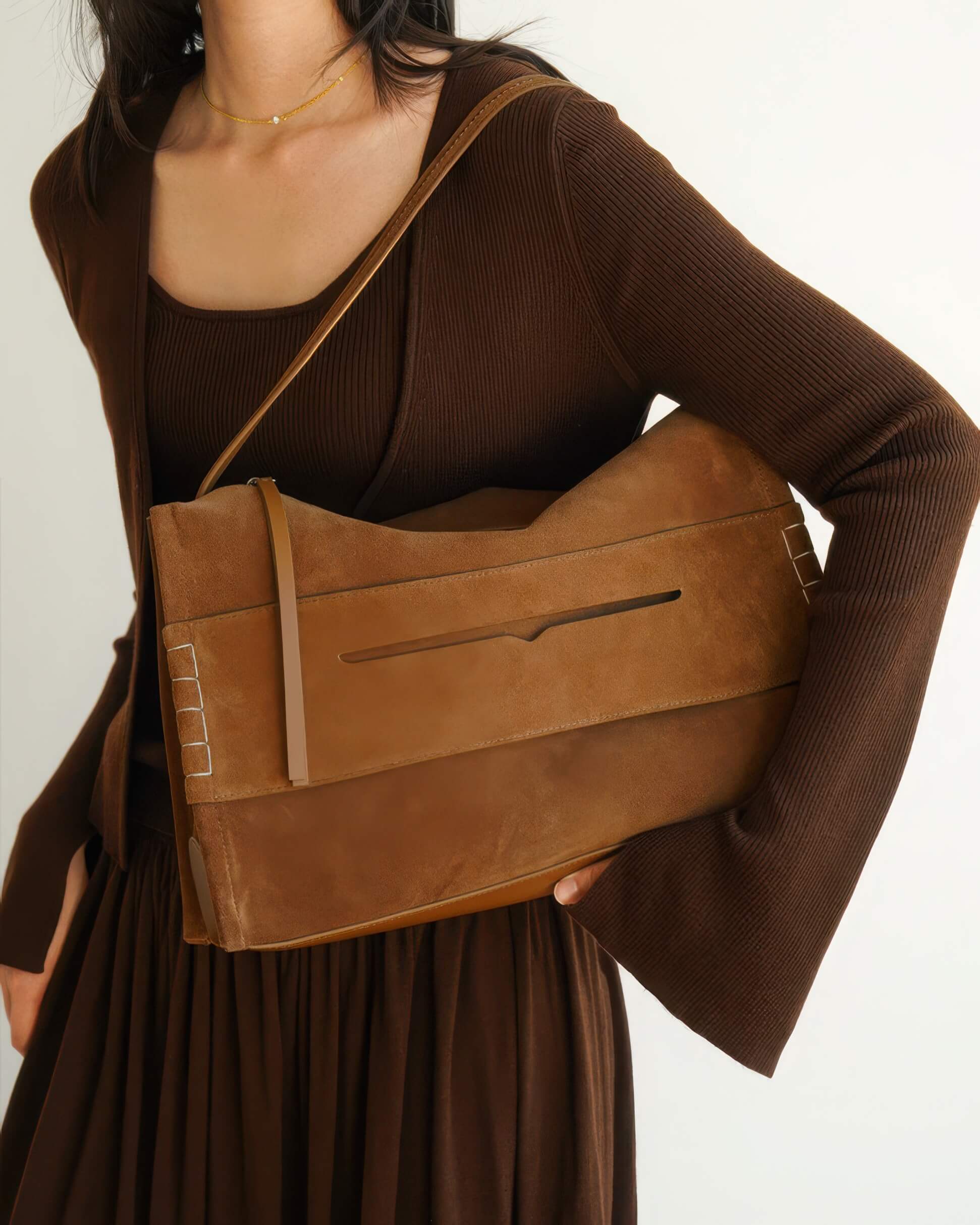 A model tucking the large brown suede shoulder bag under her arm, demonstrating its use as an oversized clutch or shoulder bag.