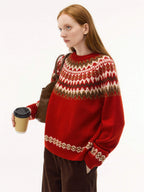 A medium shot of the model wearing a vibrant red version of the Nordic sweater, featuring white, brown, and tan jacquard patterns. She is carrying a brown leather bag and a coffee cup, styled with dark brown corduroy trousers.