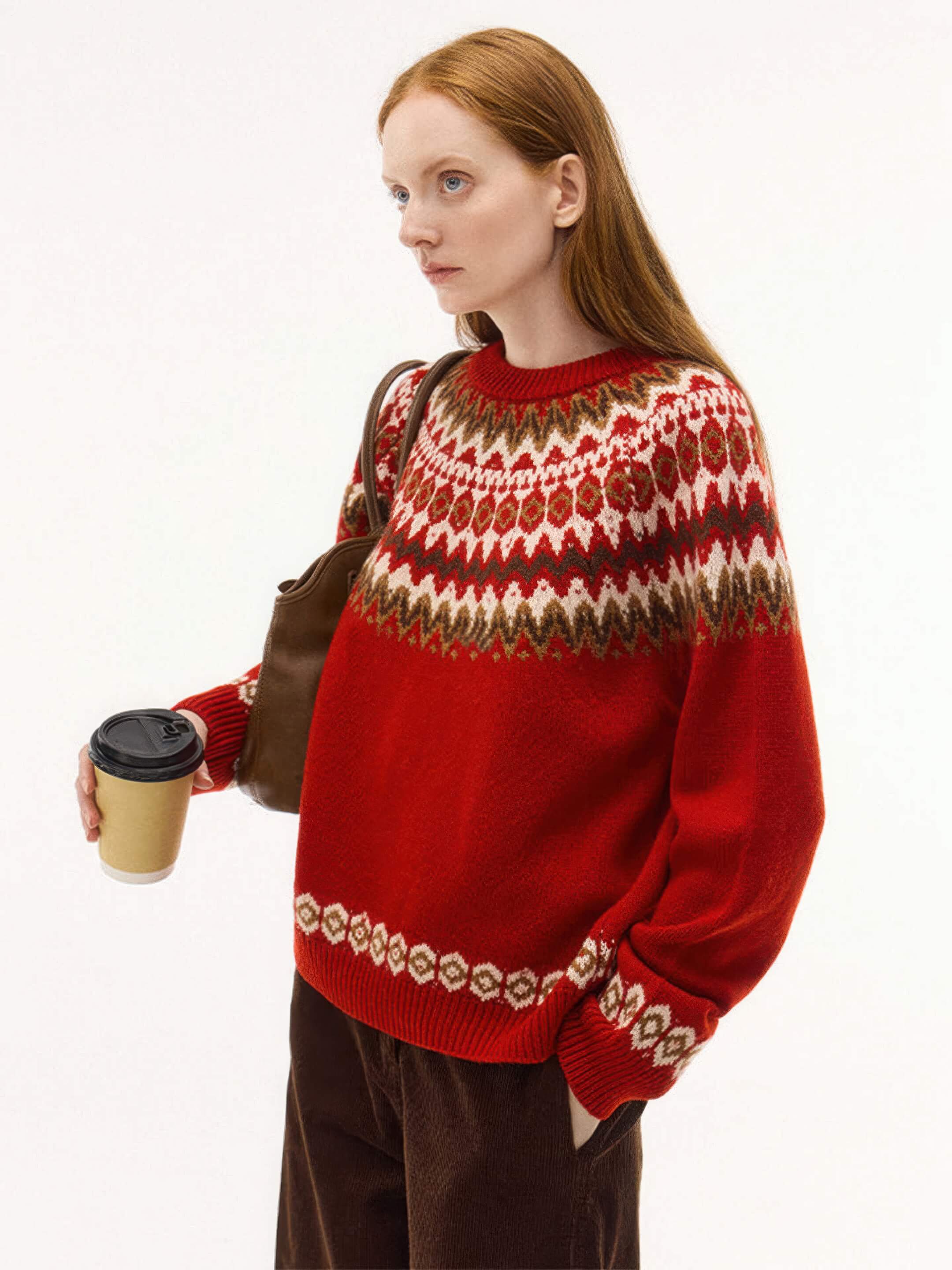 A medium shot of the model wearing a vibrant red version of the Nordic sweater, featuring white, brown, and tan jacquard patterns. She is carrying a brown leather bag and a coffee cup, styled with dark brown corduroy trousers.