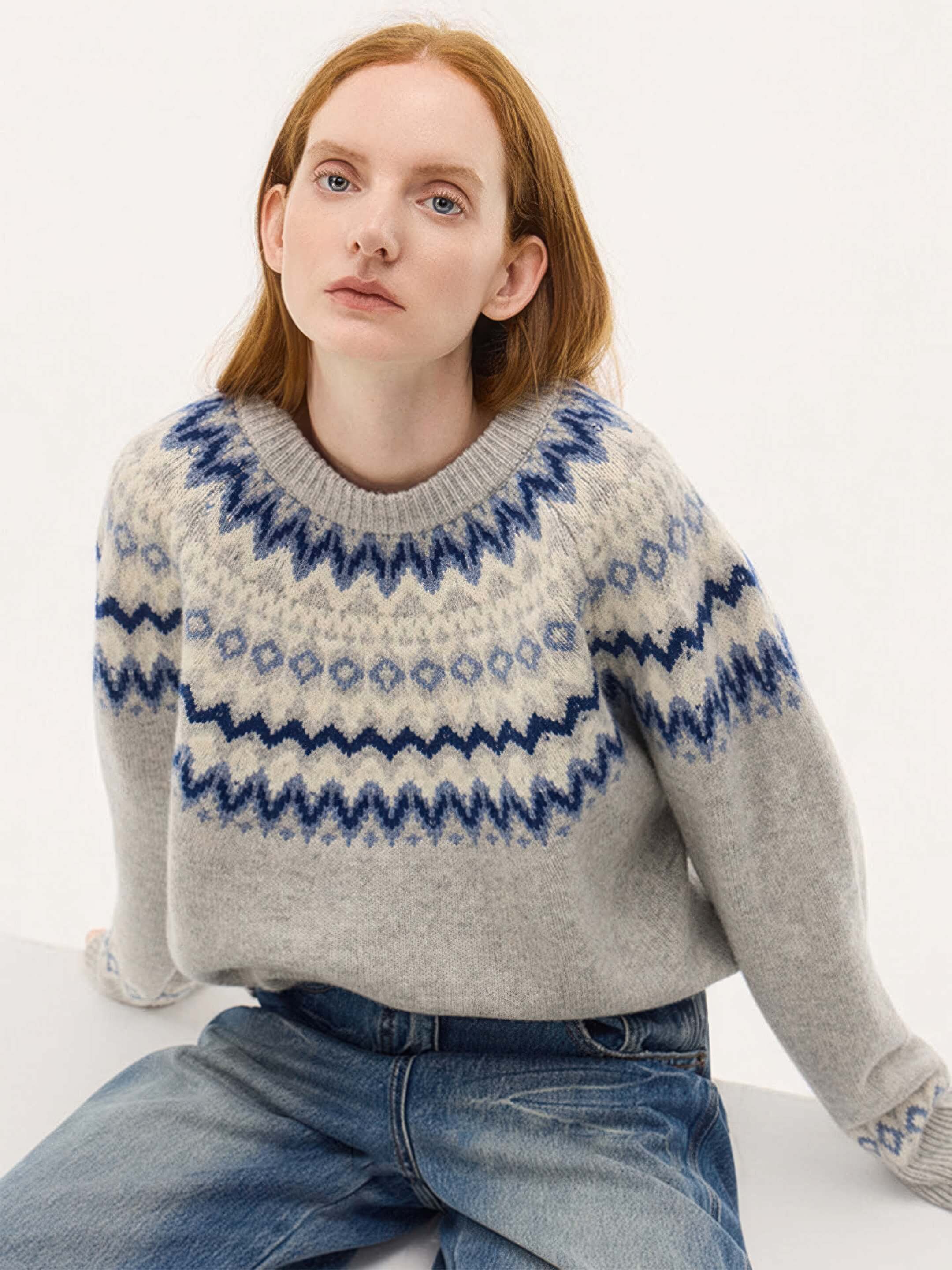 A lifestyle shot of the model sitting, showcasing the crew neckline and the intricate diamond and zigzag jacquard details of the grey and blue sweater.