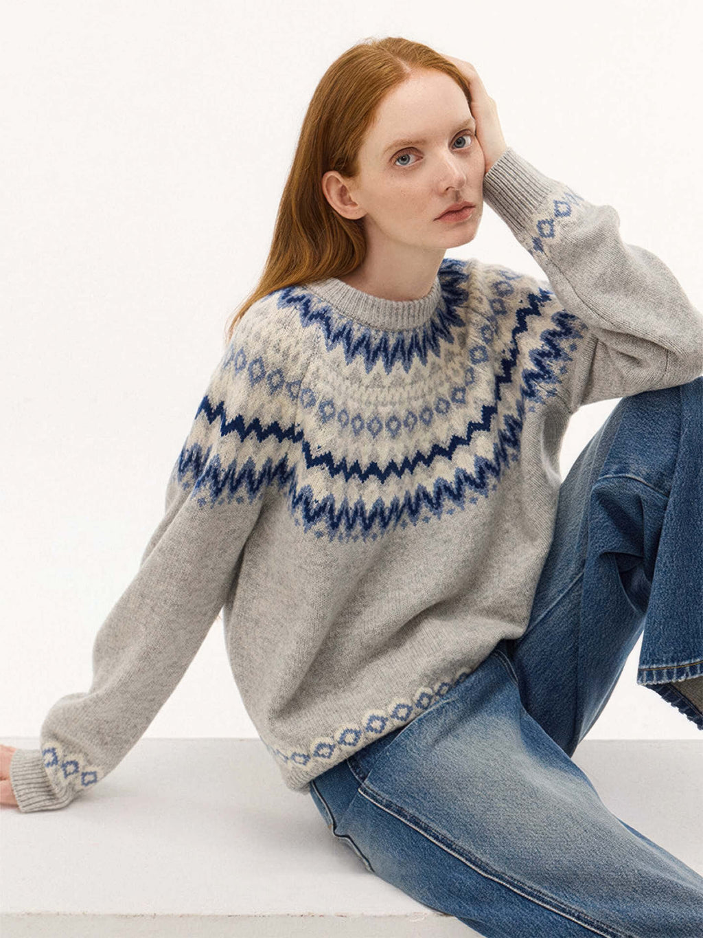 The model is seated and leaning on one hand, providing a clear view of the blue patterned sleeve cuffs and the soft, fuzzy texture of the grey wool-blend fabric.