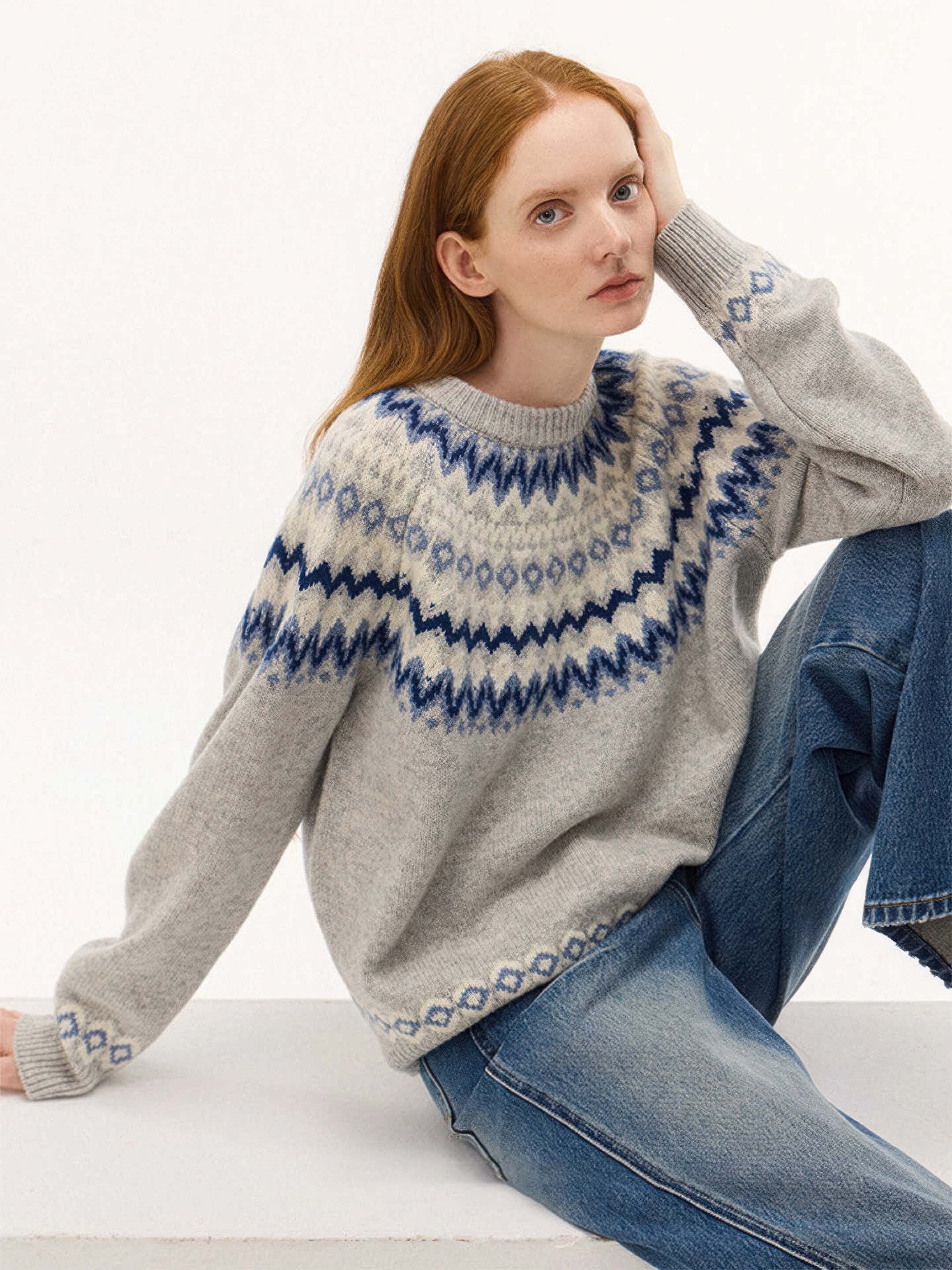The model is seated and leaning on one hand, providing a clear view of the blue patterned sleeve cuffs and the soft, fuzzy texture of the grey wool-blend fabric.
