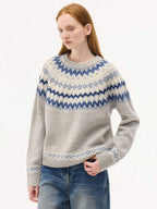 A front-facing view of a model wearing a light grey jacquard knit sweater with blue and navy Nordic patterns around the yoke, hem, and cuffs. She is paired with wide-leg denim jeans.