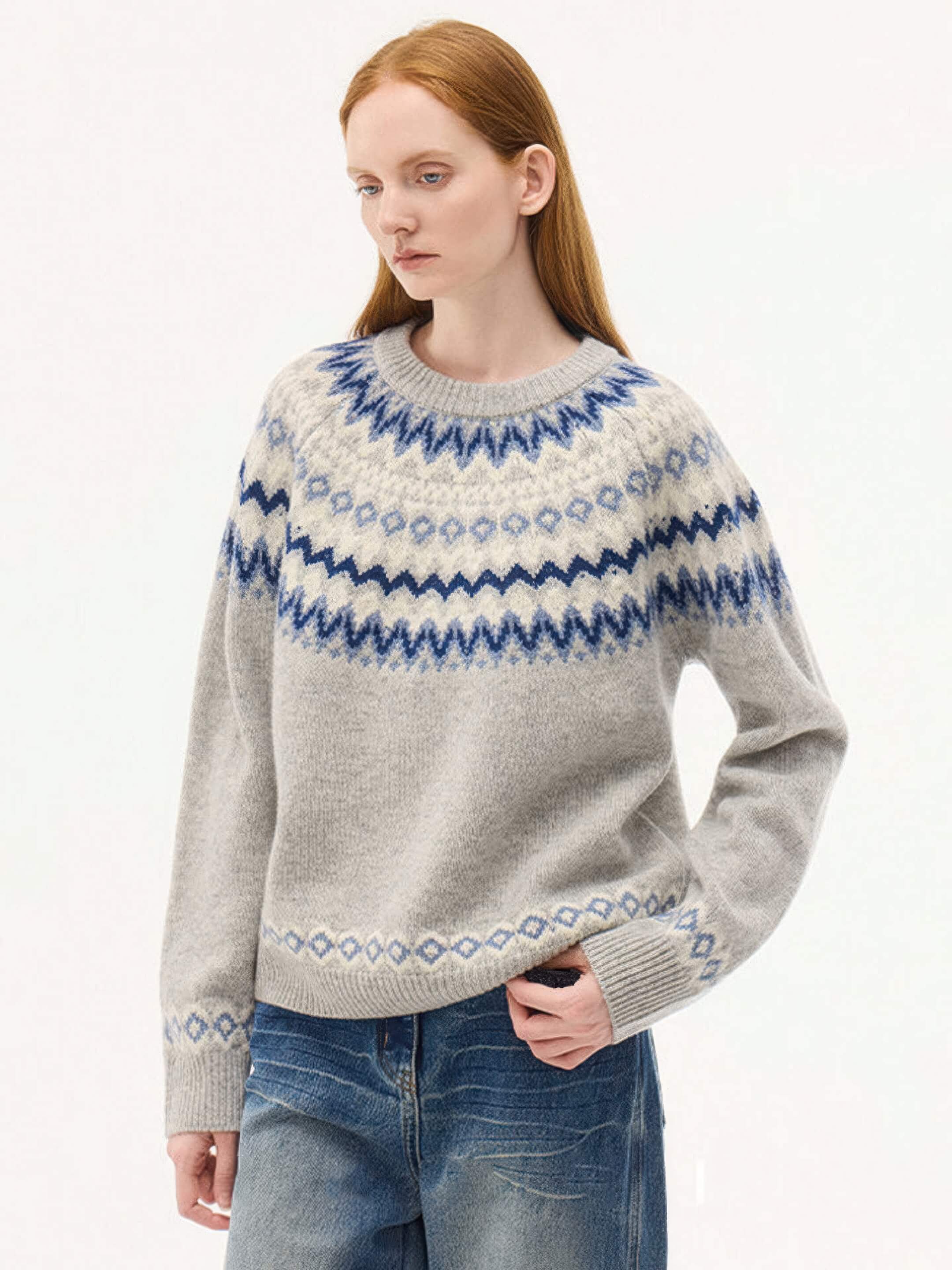 A front-facing view of a model wearing a light grey jacquard knit sweater with blue and navy Nordic patterns around the yoke, hem, and cuffs. She is paired with wide-leg denim jeans.