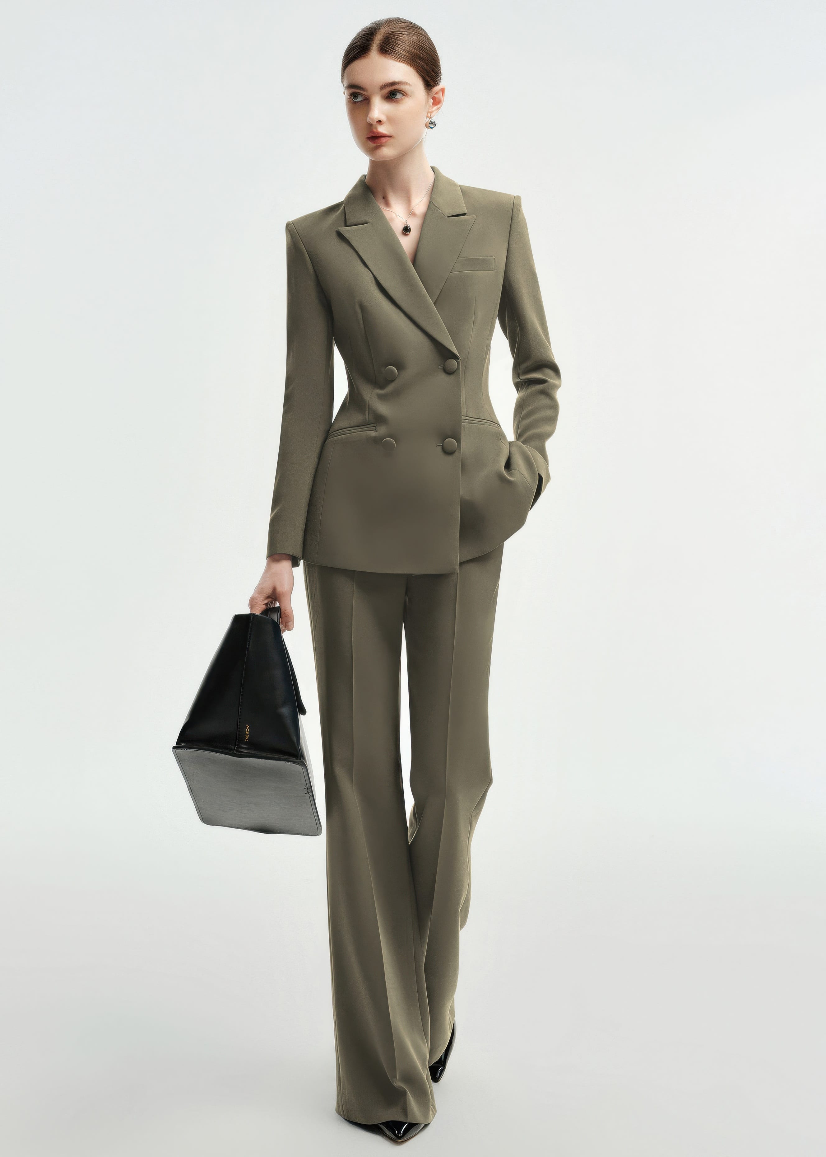 A woman walking forward in an olive green double-breasted tailored pantsuit, carrying a black leather tote bag in her right hand.