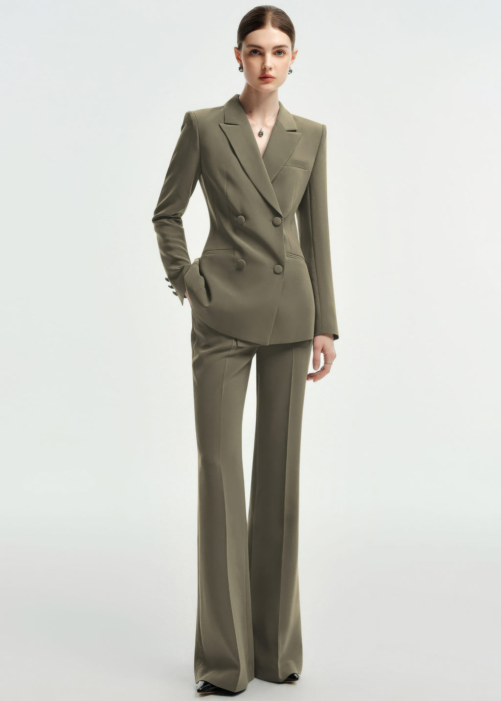 A full-body shot of a woman in an olive green double-breasted tailored pantsuit, standing against a plain white background.