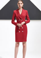 A woman standing with her hands clasped, wearing a red shawl lapel blazer dress with white button detailing.