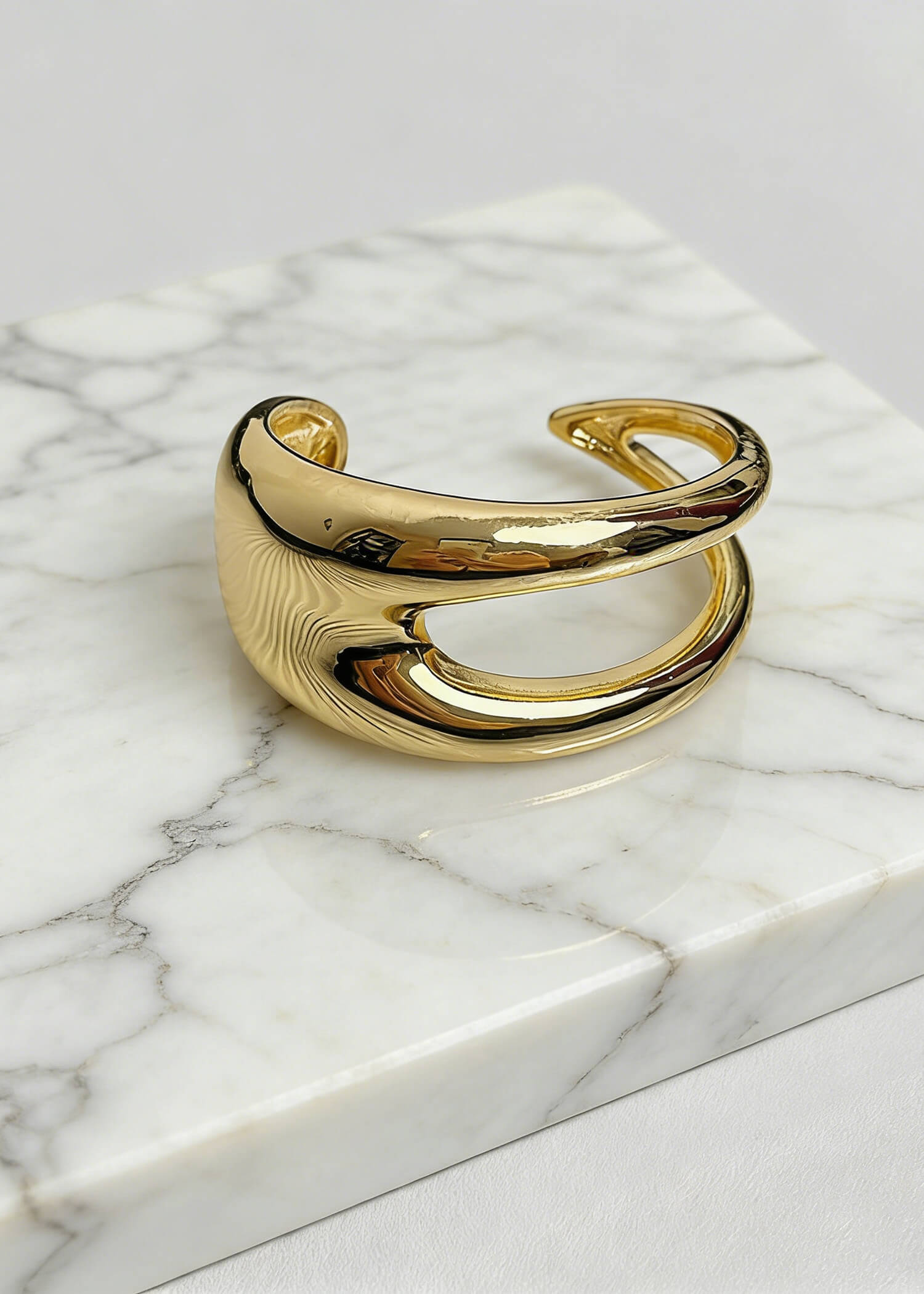 Product photography of a gold sculptural hollow cuff metal bracelet displayed on a marble block to emphasize its luxury aesthetic.