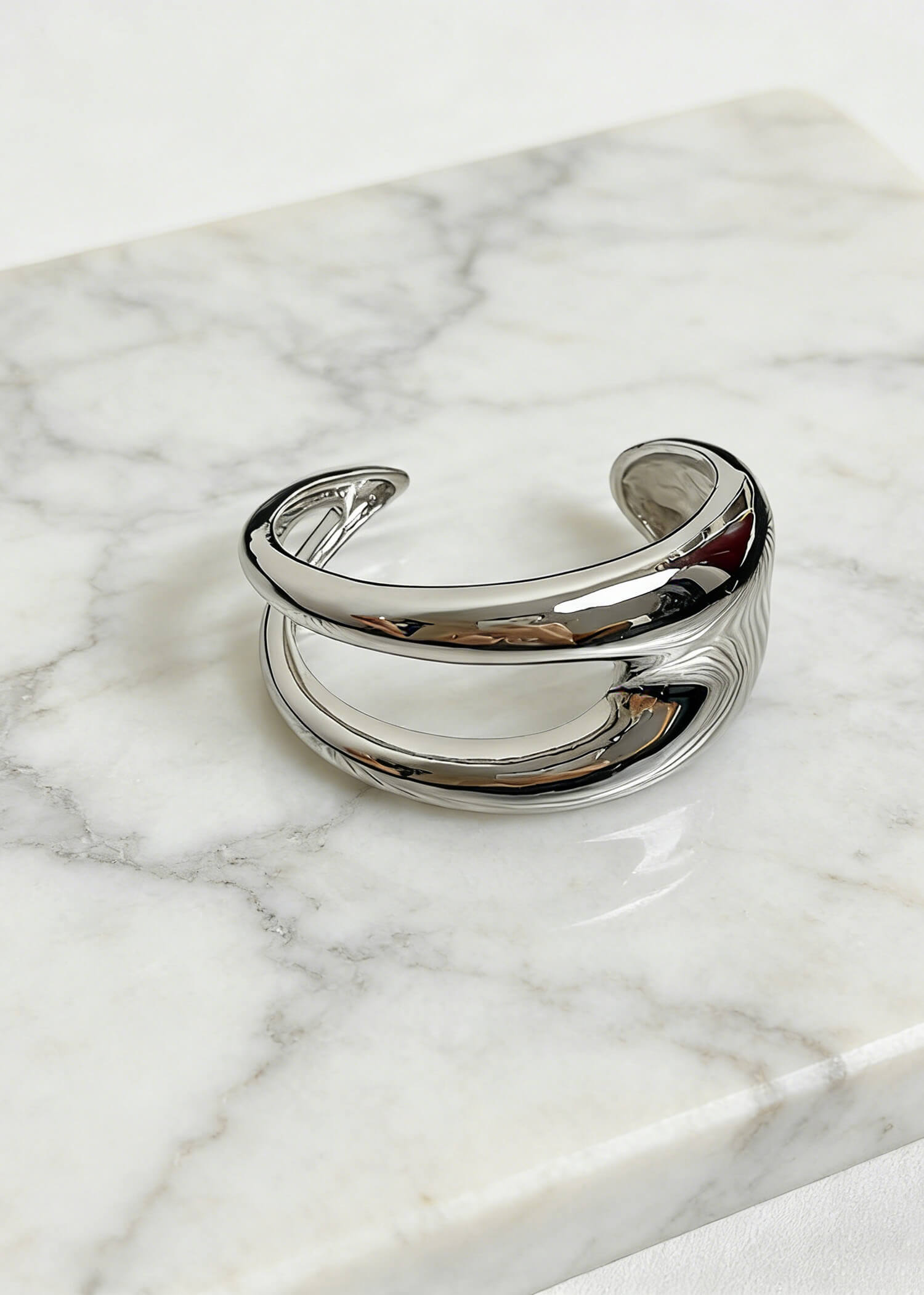 Still life of a silver sculptural hollow cuff metal bracelet resting on a white marble surface to show texture and polish.