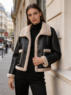 Model leaning against a stone building wearing a black leather aviator jacket with beige shearling trim