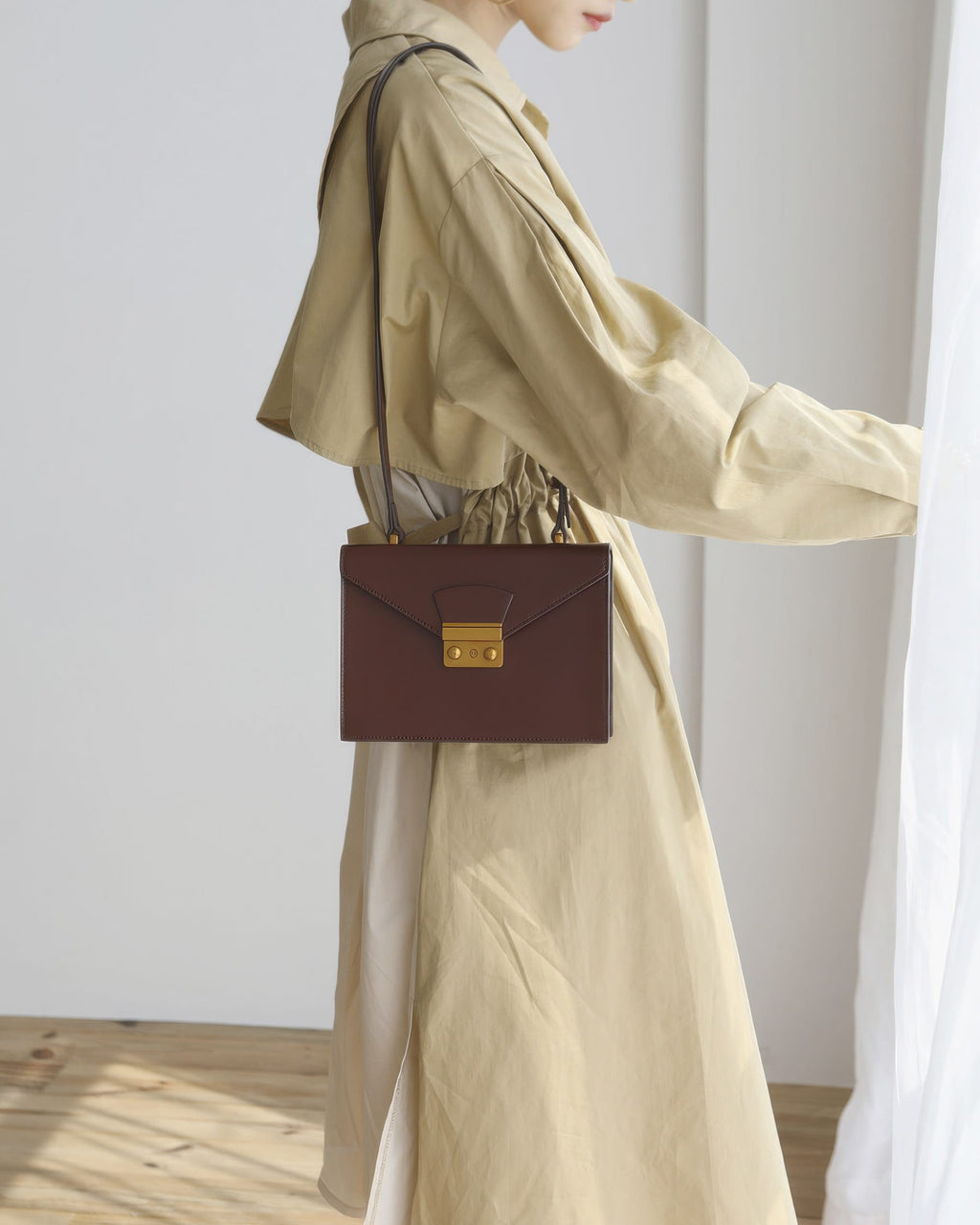 Side profile shot of a model wearing a beige dress standing by a curtain while carrying a dark brown structured leather envelope flap shoulder bag.
