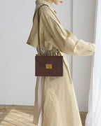 Side profile shot of a model wearing a beige dress standing by a curtain while carrying a dark brown structured leather envelope flap shoulder bag.