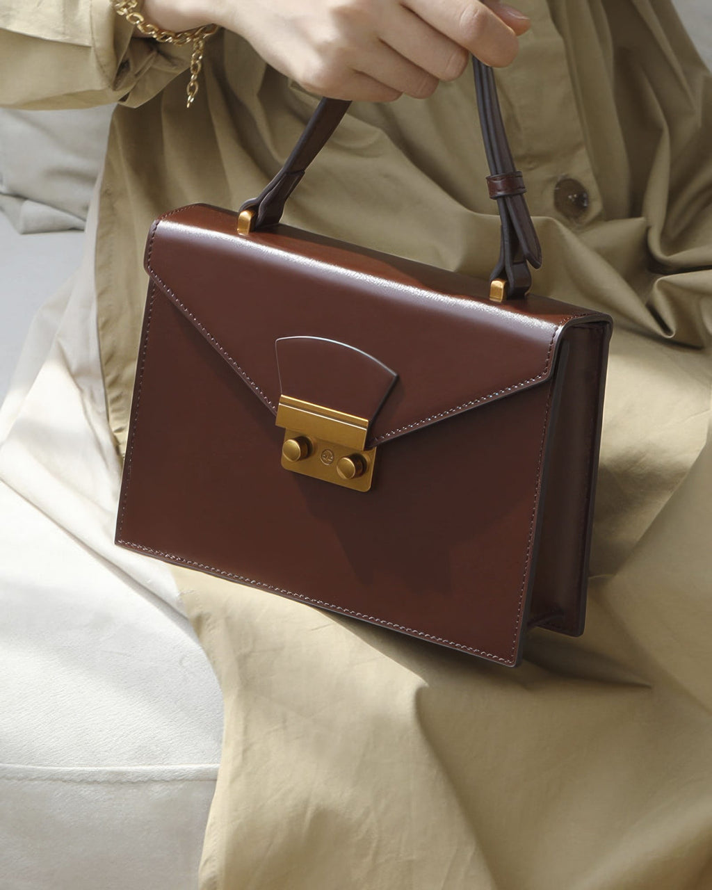 Close-up view of a chestnut brown leather envelope flap shoulder bag with gold hardware held by a model wearing a beige dress.