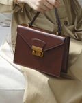 Close-up view of a chestnut brown leather envelope flap shoulder bag with gold hardware held by a model wearing a beige dress.