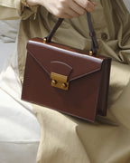 Close-up view of a chestnut brown leather envelope flap shoulder bag with gold hardware held by a model wearing a beige dress.