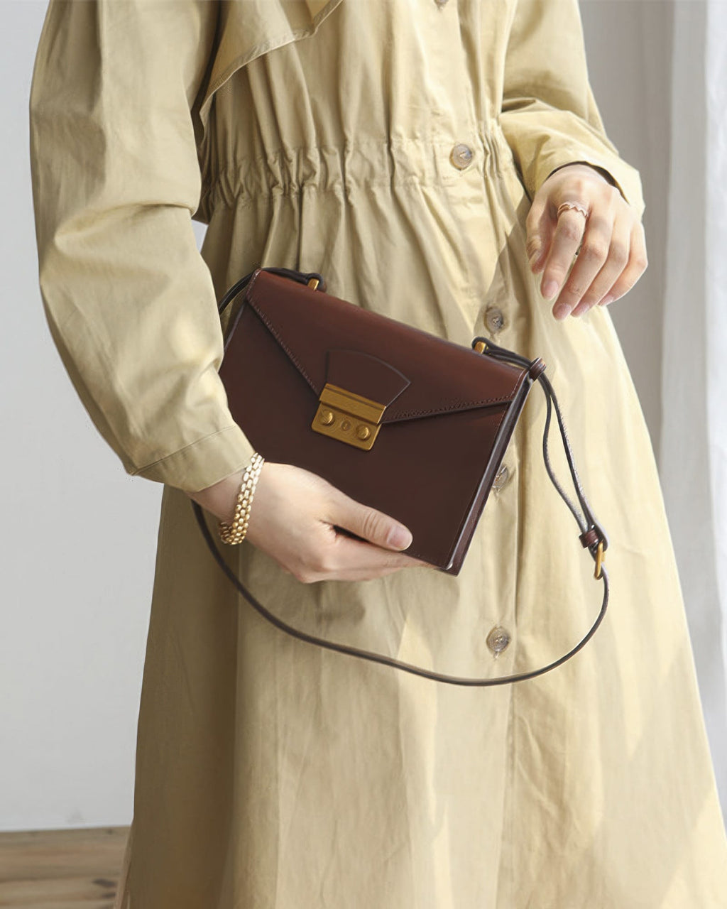 Mid-section shot of a model in a beige trench dress holding a dark brown structured leather envelope clutch with gold clasp hardware.
