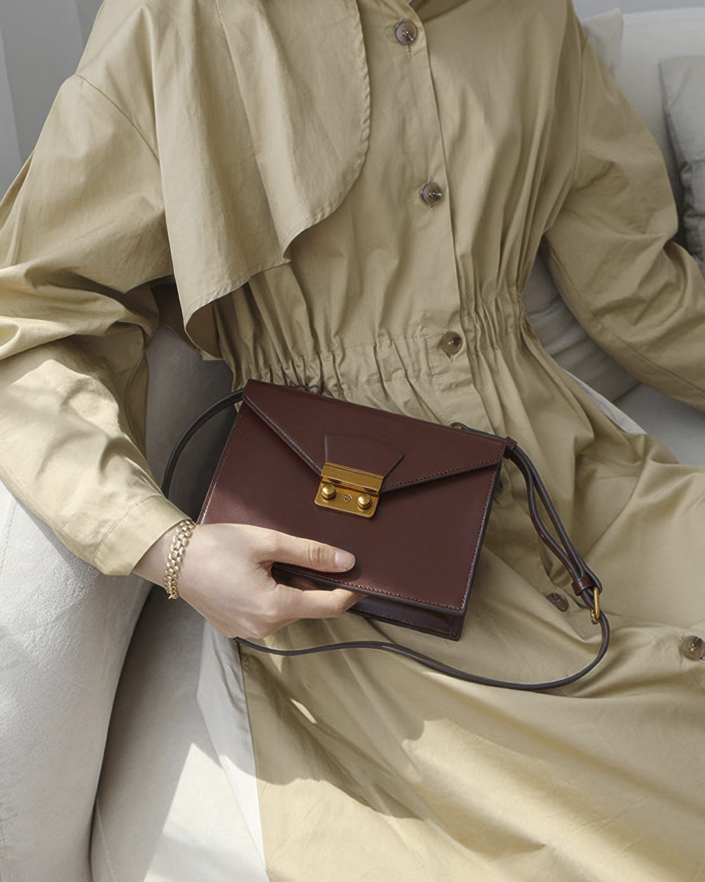 Model sitting on a couch wearing a beige trench dress holding a dark brown leather envelope flap shoulder bag.